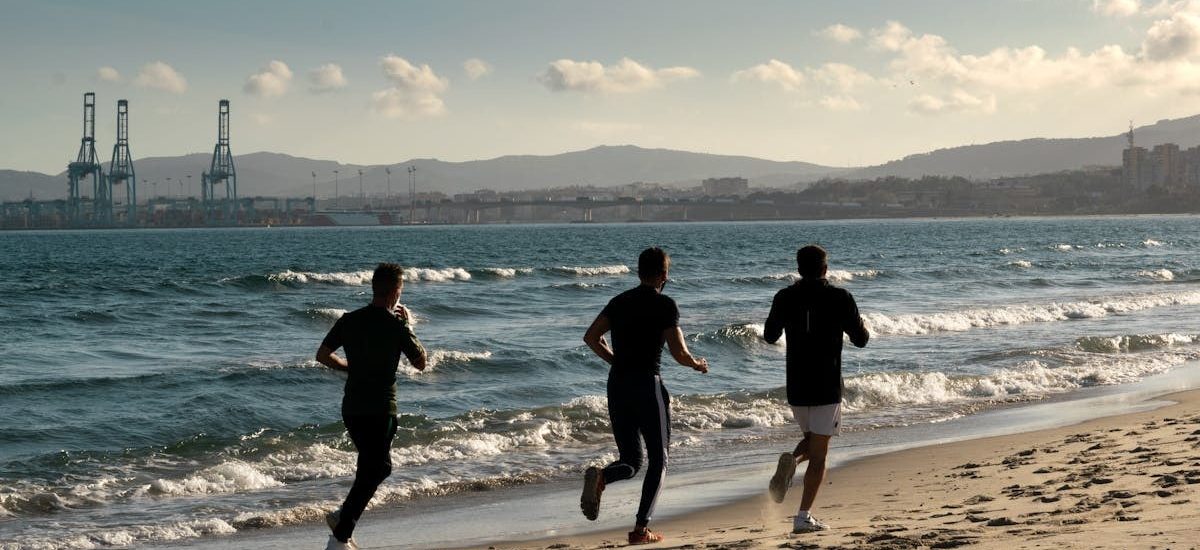 Three men jogging along a scenic beach, embracing a healthy lifestyle under a clear sky.